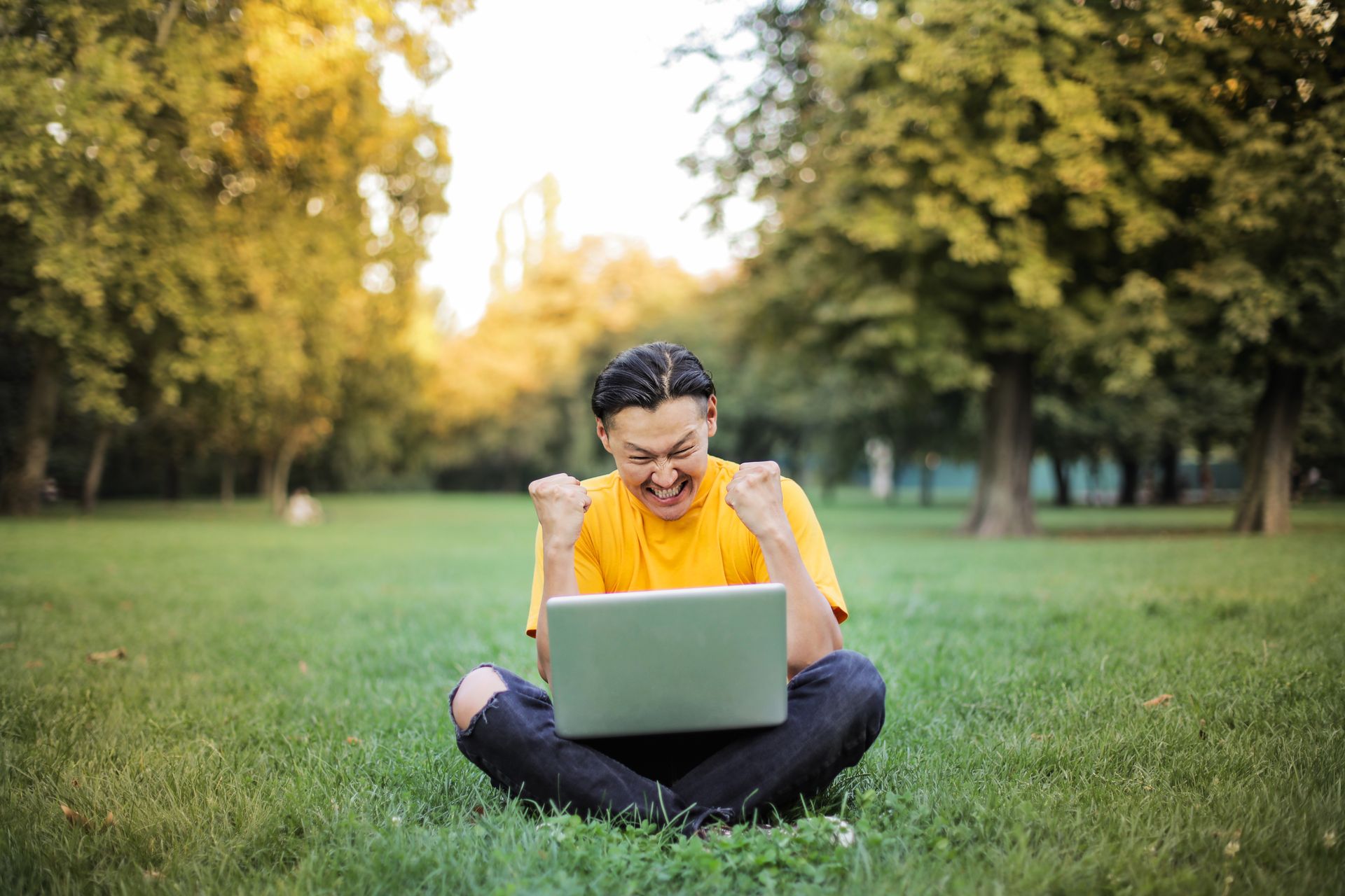 A student in a yellow shirt laughing and pumping both fists at his laptop in a sunlit park after seeing the SAT practice score he was aiming for